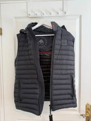 Svart dunväst från Moose Knuckles - Snygg svart dunväst från Moose Knuckles med quiltad design och två dragkedjeförsedda fickor. Västen har hög krage, avtagbar huva och diskret logotyp på bröstet. Perfekt för lager-på-lager och håller dig varm under kyliga dagar.