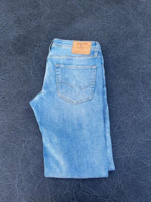 Ljusblå slim fit jeans Jack & Jones - Snygga ljusblå slim fit jeans från Jack & Jones med klassisk femficksdesign och läderpatch bak i midjan. Jeansen har raka ben och är tillverkade i mjuk denim med lätt tvättad look. Perfekta för en avslappnad stil. Modell slim Glenn storlek W32 L32