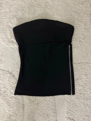 Svart tubtopp med dragkedja - Snygg svart tubtopp med en silverfärgad dragkedja längs sidan. Toppen är axelbandslös och har en stilren, tight passform som framhäver figuren. Perfekt att matcha med höga jeans eller kjol för en trendig look.