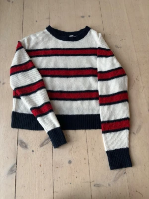 Randig stickad tröja i vitt, rött och svart - Snygg stickad tröja med breda blåa och röda ränder på vit botten. Tröjan har rund halsringning, ribbstickade muddar och avslut. Perfekt för dig som gillar en klassisk men trendig look.