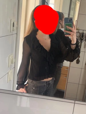 Svart volangblus från Zara - Trendig svart blus från Zara med transparenta, långärmade ärmar och volangdetaljer längs den djupa v-ringningen. Blusen är croppad och har en lätt rynkad struktur i tyget. Perfekt för dig som gillar en chic och feminin stil.
