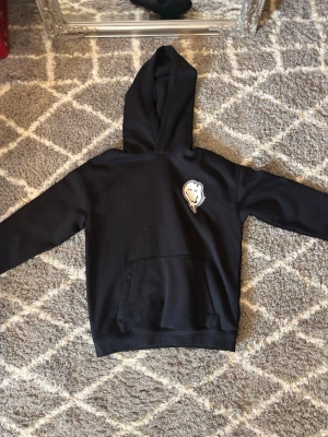 Svart hoodie med tryck och huva - Svart hoodie med stor huva och magficka. På bröstet finns ett vitt tryck av ett smältande leende ansikte och på ryggen står det 'NOTHING IS REAL BUT NOW' i stor, vågig text. Perfekt för dig som gillar streetwear och unika prints.