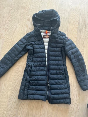 Svart lång dunjacka från Parajumpers - Säljer en svart lång dunjacka från Parajumpers med huva och snygga detaljer. Jackan har quiltad design, dragkedja framtill och ett coolt PJS-märke på ärmen. Perfekt för kalla vinterdagar och riktigt stilren att bära.