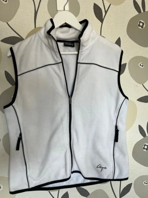 Vit fleeceväst från Clique - Vit fleeceväst från Clique med svarta detaljer och dragkedja framtill. Västen har hög krage, svarta kantband och två fickor med dragkedja. Perfekt för lager-på-lager och enkel att matcha till olika outfits. Storlek M men lång på mig (186)