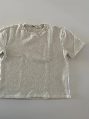 Vit kortärmad t-shirt i frotté - Säljer en stilren vit t-shirt i mjuk frotté med rund halsringning och korta ärmar. Perfekt för dig som gillar minimalistisk och clean stil. Passformen är normal och materialet känns riktigt skönt mot huden. Storlek S men passar mig som xs. Kortare i modellen
