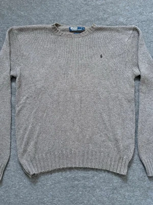 Beige stickad tröja från Polo Ralph Lauren - Denna klassiska Polo Ralph Lauren-tröja är den perfekta kombinationen av komfort och stil. Den är stickad i en mjuk, grå nyans som passar till allt, och har en avslappnad passform som är perfekt för vardagsbruk. Polo-hästlogotypen är subtilt placerad på bröstet, vilket ger en touch av lyx. Oavsett om du klär upp dig eller klär ner dig, kommer denna tröja garanterat att bli ett favoritalternativ. Finns inga märken eller fläckar. Skickas inom 1-2 dagar:)