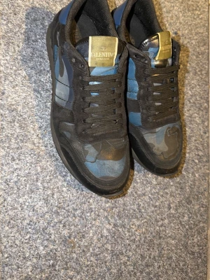 Valentino rockrunners camo sneakers i blått och svart - Snygga sneakers från Valentino med blå och svarta camo-detaljer. Skorna har svarta snören, guldiga logotaggar på plösen och en klassisk låg siluett. Materialmix av mocka och textil ger en cool och exklusiv känsla. Skick 8/10 Lite små defekter men inget stort, se själv på bilder. Hör av dig vid frågor eller fler bilder. Självklart äkta!!!