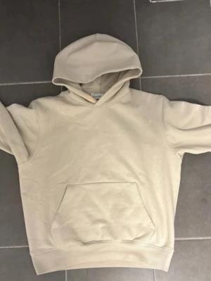 Beige hoodie med magficka - Snygg beige hoodie med klassisk huva och stor magficka framtill. Hoodien har ribbade muddar vid ärmslut och nederkant. Tillverkad i mjukt bomullsmaterial som känns skönt mot huden. Perfekt för en avslappnad och trendig look.