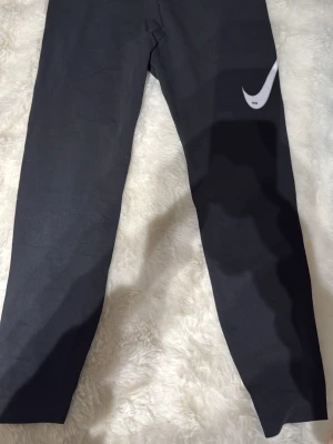 Svarta leggings från Nike - Snygga svarta leggings från Nike med klassisk vit Swoosh-logga på ena benet. Tight passform och stretchigt material som sitter skönt. Perfekta till träning eller chill outfits. Materialet känns mjukt och följsamt.