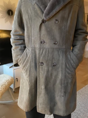 Grå mockakappa med pälskrage - Stilren grå mockakappa med dubbelknäppning och bred pälskrage. Kappan har två snedställda fickor framtill och en rak passform som går ner över låren. Det är äkta mocka.                                                        Jag som modell är 183 lång, ungefär L