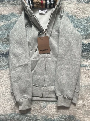 Grå burberry zip - Grå zip hoodie från Burberry med klassiskt rutigt foder i huvan och diskret logga broderad på bröstet. Skulle säga att det är barnstorlek på denna, 164. Pris går alltid att diskuteras 🙏