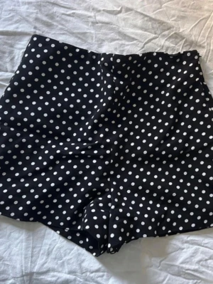 Svarta prickiga högmidjade shorts - Svarta shorts med vita prickar och hög midja. Klassisk och trendig look med en figurnära passform. Perfekta för varma dagar och lätta att matcha med olika toppar. Materialet känns mjukt och svalt mot huden.