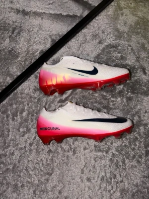 Nike Mercurial vita fotbollsskor elite - Säljer ett par Nike Mercurial fotbollsskor i vitt med rosa och röd gradient på sulan. Skorna har en svart Nike-swoosh på sidan och är tillverkade i ett lätt syntetmaterial för bästa känsla och grepp på planen. Perfekta för dig som vill sticka ut på matchen.