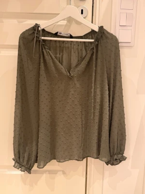 Olivgrön blus med struktur från ZARA - Olivgrön blus från ZARA med lång ärm, lätt transparent tyg och små prickar i strukturen. Blusen har v-ringning med knytband vid halsen och volangdetaljer vid ärmslut. Perfekt för en trendig och avslappnad look.