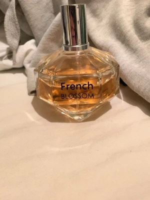 French Blossom parfym - En elegant parfym med namnet French Blossom i en multifacetterad glasflaska med silvrig spraykork. Flaskan har en ljus bärnstensfärgad vätska och en modern, stilren design som passar perfekt på sminkbordet.