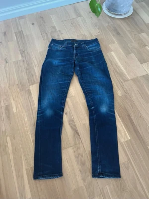 Nudie Jeans - Snygga mörkblå jeans från Nudie Jeans med klassisk femficksmodell och raka ben. Jeansen har slitningar och ljusare partier framtill för en cool, använd look. Tillverkade i denim med tydliga kontrastsömmar och knappgylf.