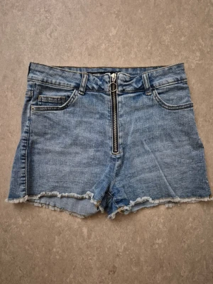 Jeansshorts - Säljer ett par blå jeansshorts med råa kanter och en cool dragkedja framtill. Shortsen har klassiska fickor både fram och bak samt hällor för bälte. Perfekta för varma dagar och ger en avslappnad vibe.