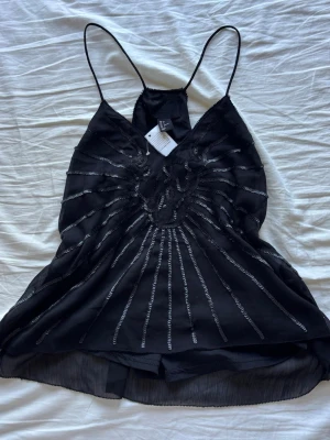 Svart paljettlinne från H&M - Supersnyggt svart linne från H&M med tunna axelband och glittriga paljettdetaljer i ett strålande mönster framtill. 