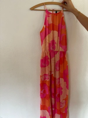 Färgglad långklänning från Bik Bok - Snygg långklänning från Bik Bok med stora blommiga mönster i rosa, orange och beige toner. Klänningen har smala axelband och en luftig passform, perfekt för varma dagar. Tillverkad i ett lätt och skönt polyestertyg.