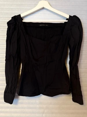Svart långärmad blus från Zara - Trendig svart blus från Zara med markerad midja och fyrkantig urringning. Blusen har långa puffärmar och är tillverkad i ett lätt, vävt tyg som ger en snygg siluett. Perfekt att styla med både jeans och kjol.