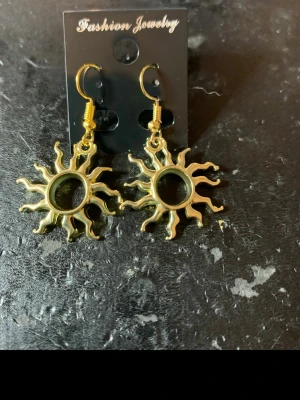 Solformade guldörhängen - Ett par coola örhängen i form av solar, tillverkade i guldfärgad metall. De har en unik och lekfull design med strålande solstrålar och är perfekta för att ge din stil en extra edge. Hängande modell med krokfäste.