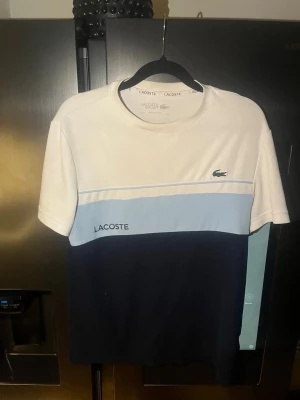 Lacoste t-shirt med färgblock - Snygg t-shirt från Lacoste med färgblock i vitt, ljusblått och mörkblått. Klassisk krokodillogga på bröstet och Lacoste-tryck. Rund halsringning och korta ärmar. Perfekt för en stilren och sportig look.