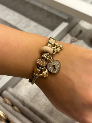 Armband med berlocker - Snyggt armband i silverfärg med flera berlocker i guld- och roséguldtoner. Armbandet har detaljer som hjärtan, lås och pärlor, samt en hjärtberlock med glittrande stenar. Perfekt accessoar för att lyfta din stil. Köpt i marknad i Spanien.