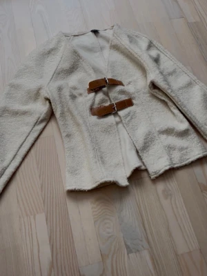 Krämvit cardigan med spännen - Stilren krämvit cardiganjacka med två bruna bältespännen framtill. Jackan har lång ärm och är gjord i ett mjukt, strukturerat tyg som ger en mysig känsla. Perfekt för lager-på-lager och en cool vardagslook.