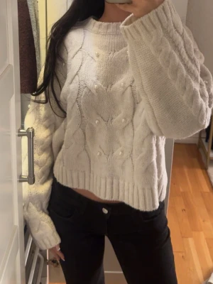 Vit kabelstickad tröja med pärlor - Superfin vit kabelstickad tröja med pärldetaljer på framsidan. Tröjan har en boxy, croppad passform och breda ärmar med ribbade muddar. Perfekt för att hålla sig varm och snygg under kyliga dagar.