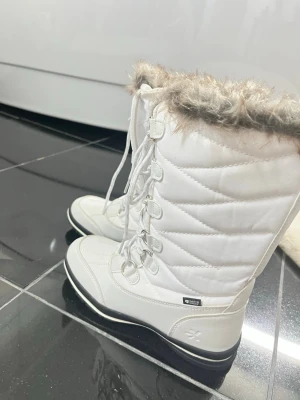 Vita vinterstövlar med fuskpäls - Säljer ett par vita vinterstövlar från Sköna Marie med fuskpäls upptill och snörning framtill. Stövlarna har en robust svart sula med mönster för extra grepp och är tillverkade i vattenavvisande material. Perfekta för kalla dagar och snö.