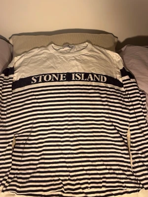 Randig långärmad tröja Stone Island - Snygg långärmad tröja från Stone Island med marinblå och vita horisontella ränder. Stor logga över bröstet och rund halsringning. Tillverkad i mjukt bomullsmaterial, perfekt för en avslappnad och trendig stil.