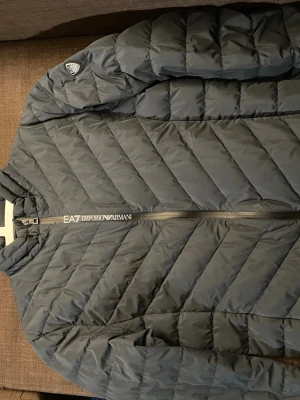 Grå dunjacka från EA7 Emporio Armani - Snygg grå dunjacka från EA7 Emporio Armani med diskret logga på bröstet och dragkedja framtill. Jackan har ett quiltat mönster i diagonala ränder och står för både stil och värme under kalla dagar. Perfekt för dig som vill ha en modern look.
