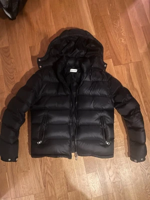 Svart pufferjacka från Moncler - Säljer en svart pufferjacka från Moncler med avtagbar huva och två dragkedjeförsedda fickor framtill. Jackan har en klassisk quiltad design och blanka tryckknappar vid ärmsluten. Perfekt för kalla vinterdagar och riktigt snygg streetstil.