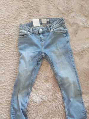 Ljusblå skinny jeans från 157 - Säljer ett par ljusblå skinny jeans från 157 med klassisk femficksmodell och normalhög midja. Jeansen har bälteshällor, knappgylf och är tillverkade i mjukt denimtyg med lite stretch för extra komfort. Perfekta för en avslappnad och trendig look.