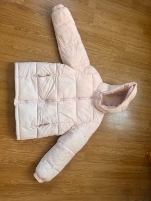 Ljusrosa pufferjacka med huva - Säljer en ljusrosa pufferjacka med dragkedja och två sidofickor. Jackan har en stor, mysig huva med dragsko och quiltad design. Perfekt för kalla dagar och riktigt skön att bära. Materialet är syntet och jackan är vadderad för extra värme.