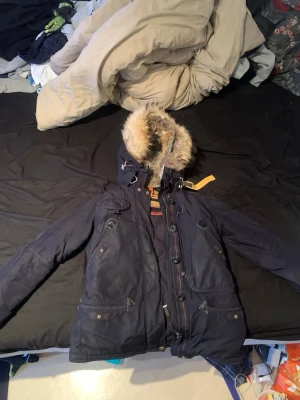 Mörkblå dunjacka från Parajumpers - Snygg mörkblå dunjacka från Parajumpers med avtagbar huva och fluffig fuskpäls. Jackan har flera fickor, dragkedja och knappar framtill samt den klassiska gula detaljen vid halsen. Perfekt för kalla dagar och riktigt varm.