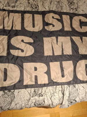 Svart tygflagga med text - Stor svart tygflagga med stora vita bokstäver som säger 'MUSIC IS MY DRUG'. Perfekt som väggdekoration för musikälskare eller till festivaler och events. Tillverkad i tunt tygmaterial och lätt att hänga upp.