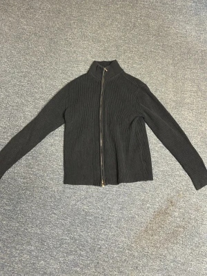 Svart ribbstickad ziptröja - Svart ribbstickad tröja med dragkedja framtill och hög krage. Långärmad och i ett mjukt, stretchigt material som ger en clean och stilren look. Perfekt för lager-på-lager och enkel att matcha med andra plagg.