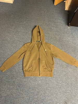 Olivgrön hoodie från Polo Ralph Lauren - Snygg olivgrön hoodie med dragkedja från Polo Ralph Lauren. Klassisk huva med dragsko, två fickor framtill och broderad vit logga på bröstet. Perfekt för en avslappnad och stilren look.