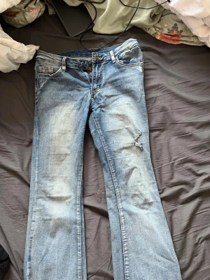 Blå bootcut jeans med slitning - Säljer ett par blå bootcut jeans med slitningar/snygg detalj. Jeansen har klassisk femficksdesign och ljus tvätt med snygga detaljer. Perfekt för dig som gillar en avslappnad och trendig look. Det är ett litet hål vid ena innerbenet men det är lätt att sy ihop.