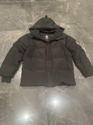 Svart pufferjacka från Canada Goose - (Pris kan självklart diskuteras😊) Säljer min svarta pufferjacka från Canada Goose eftersom den är lite stor på mig. Med huva och klassisk logotyp på ärmen. Jackan har dragkedja och knappar framtill samt två stora fickor. Perfekt för kalla vinterdagar tack vare det tjocka fodret och den rejäla designen.
