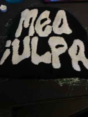 Svart mössa med texten Mea Culpa - Säljer en svart mössa med stor vit text framtill där det står 'Mea Culpa' i ett coolt, grafiskt typsnitt. Mössan är ribbstickad och har en avslappnad passform. Perfekt accessoar för dig som vill sticka ut lite extra.