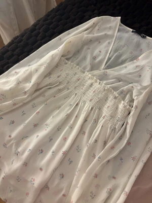 Vit blommig omlottklänning - Supersöt vit kortklänning med små blommor i rosa och blått. Klänningen har omlottliv, v-ringning, långa ärmar med resår vid handlederna och smock i midjan för en bekväm passform. Perfekt för vår och sommar!