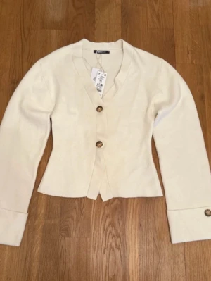 Krämvit kofta från Gina Tricot - Stilren krämvit kofta från Gina Tricot med v-ringning och två stora bruna knappar framtill. Koftan har långa ärmar med breda muddar och knappdetalj vid ärmslut. Perfekt för lager på lager och enkel att matcha med olika outfits.