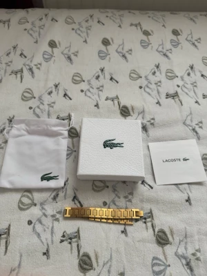 Guldigt armband från Lacoste - Snyggt guldigt armband från Lacoste med stilrena länkar och diskret Lacoste-logga på en av länkarna. Armbandet har en modern och elegant design och kommer med original ask och tygpåse. Perfekt accessoar för dig som gillar exklusiv känsla. Endast använd 1 gång, så som ny typ.