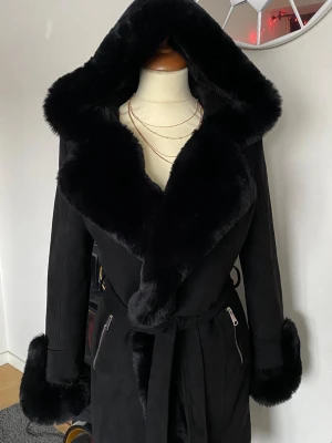 Svart kappa med fuskpäls och huva - Snygg svart kappa med stor huva och breda detaljer i mjuk fuskpäls längs krage, ärmslut och huva. Kappan har knytskärp i midjan och dragkedjefickor framtill. Perfekt för kalla dagar när du vill vara både varm och stilren.