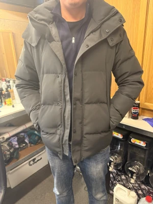 Mörkgrå pufferjacka med huva - Snygg mörkgrå canada goose pufferjacka med stor huva och dragkedja samt knappar framtill. Jackan har en quiltad design och två sidofickor. Perfekt för kalla vinterdagar och ger en clean, modern look. Materialet är troligtvis polyester. Passar L också.