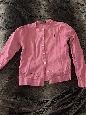 Rosa kofta från Ralph Lauren - Söt rosa kofta från Ralph Lauren med rund halsringning och vita knappar. Broderad logga på bröstet och små blommiga detaljer längs nederkanten. Perfekt för dig som gillar klassisk och feminin stil.