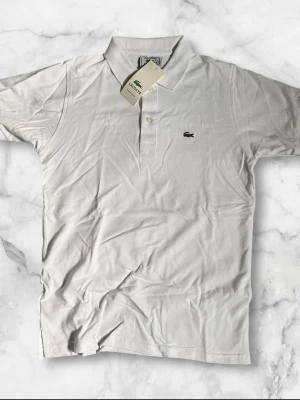 Vit pikétröja från Lacoste - Klassisk vit pikétröja från Lacoste med korta ärmar och den ikoniska krokodilloggan broderad på bröstet. Tröjan har krage och knappar framtill. Perfekt till en clean och sportig look.