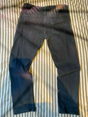 Blå jeans från Nudie Jeans - Säljer ett par klassiska blå jeans från Nudie Jeans med raka ben och snygga bakfickor med vågig söm. Jeansen har normal passform och är tillverkade i mjukt denimtyg. Perfekta för dig som gillar stilrena och tidlösa jeans.